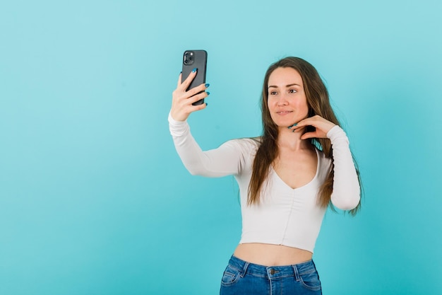 La joven se está tomando selfie con el teléfono inteligente sosteniendo la mano debajo de la barbilla en el fondo azul