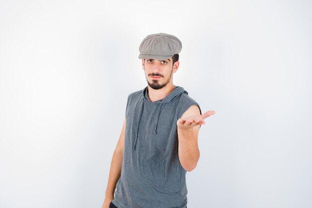 Joven estirando una mano sosteniendo algo en camiseta gris y gorra y mirando serio