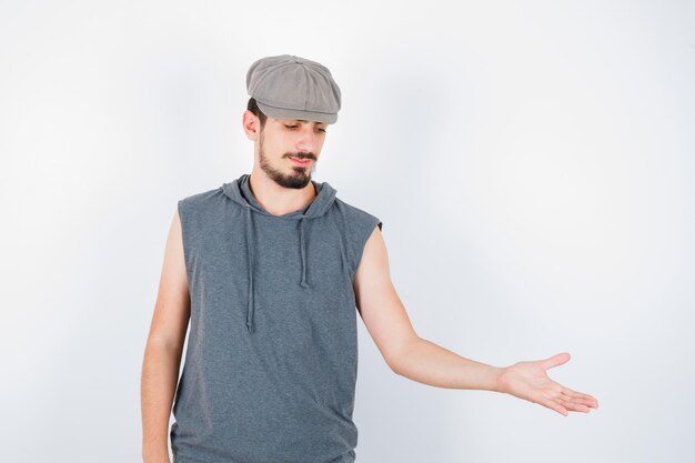 Joven estirando una mano sosteniendo algo en camiseta gris y gorra y mirando serio