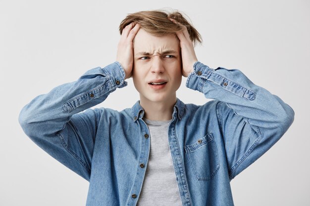 El joven estresante con las manos en el cabello rubio tiene dolor de cabeza, aprieta los dientes con dolor, vive en tensión y tiene muchos problemas. Estudiante masculino sufre de dolor, tiene expresión cansada y exhausta