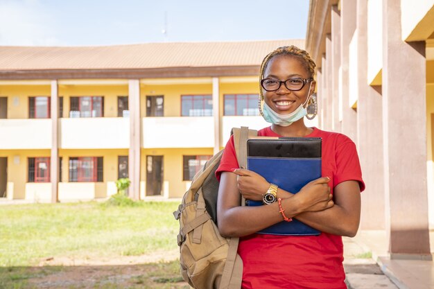 Joven estudiante africana con una mascarilla sosteniendo sus libros de texto en un área del campus