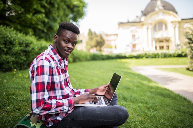Joven estudiante africano sonriente sentado en la hierba con el portátil al aire libre en verano