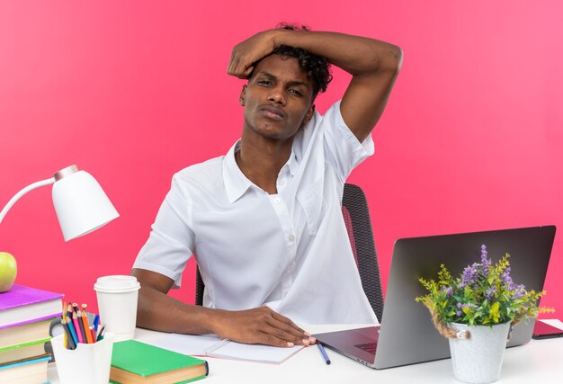 Joven estudiante afroamericano descontento sentado en el escritorio con herramientas escolares poniendo la mano en la cabeza
