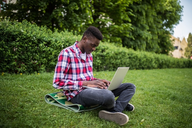 Joven estudiante afroamericano sentado en el césped con un portátil cerca del campus