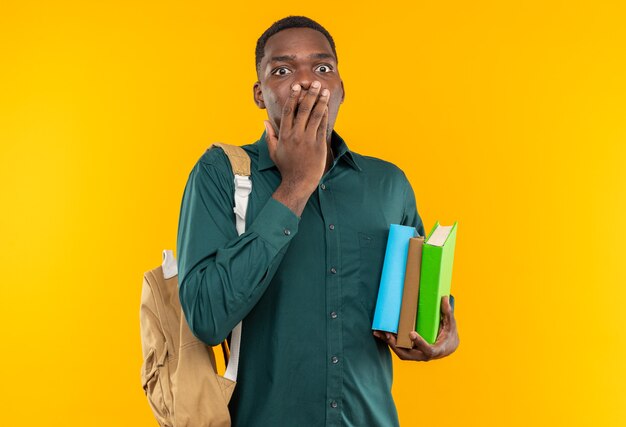 Joven estudiante afroamericano sorprendido con mochila sosteniendo libros y poniendo la mano en la boca