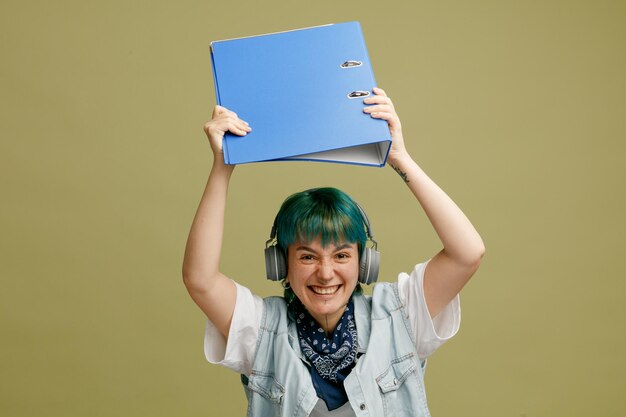 Joven estudiante agresiva con auriculares y pañuelo en el cuello levantando una carpeta mirando a la cámara preparándose para golpear a alguien con una carpeta aislada en un fondo verde oliva