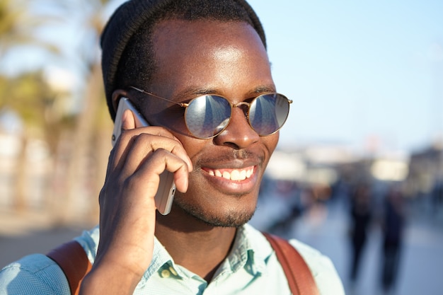 Joven estudiante alegre en gafas de sol con lentes de espejo y sombreros sonriendo felizmente