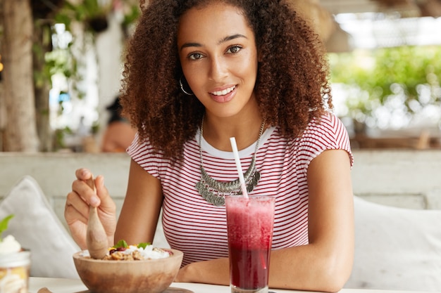 Una joven estudiante alegre y positiva que disfruta de las vacaciones de verano en el extranjero, pasa el tiempo libre en la cafetería, come ensalada y batido rojo, se viste informalmente y le gusta estar en buena compañía. Concepto de personas y estilo de vida