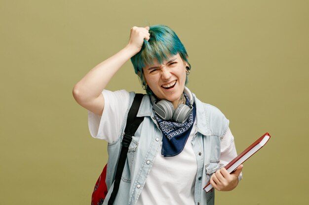 Joven estudiante arrepentida usando audífonos y pañuelo en el cuello y mochila sosteniendo un cuaderno mirando a la cámara tirando de su propio cabello aislado en un fondo verde oliva