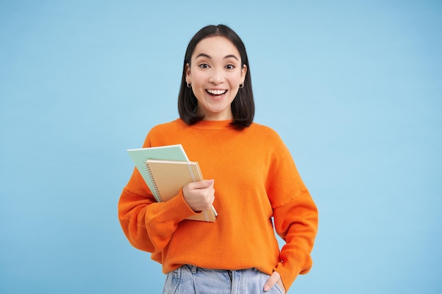 Joven estudiante asiático con cuadernos de pie en sudadera naranja mirando a la cámara posando sobre azul