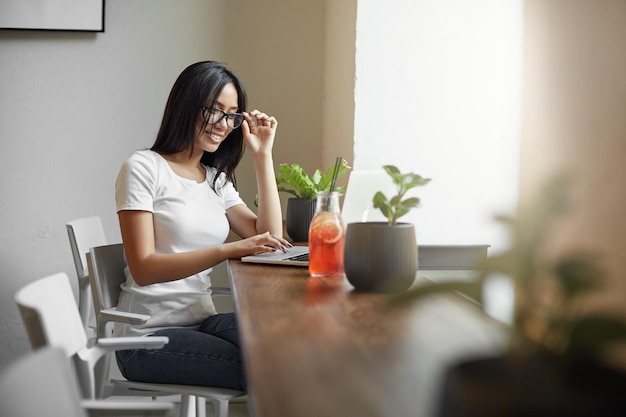 Joven estudiante asiático usando una computadora portátil en un café o coworking bebiendo limonada sentado junto a una ventana grande Concepto de estudio