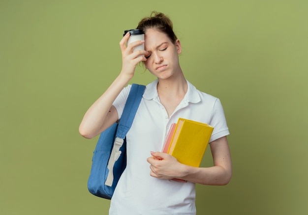 Joven estudiante bastante disgustada con bolsa trasera sosteniendo libro y bloc de notas y tocando la frente con una taza de café de plástico aislada sobre fondo verde con espacio de copia