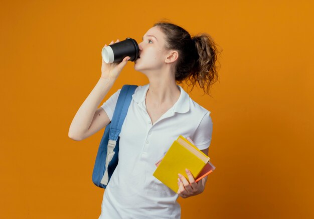 Joven estudiante bastante femenina con bolsa trasera mirando al lado sosteniendo el bolígrafo de bloc de notas de libro y tomando café de una taza de café de plástico aislada sobre fondo naranja con espacio de copia