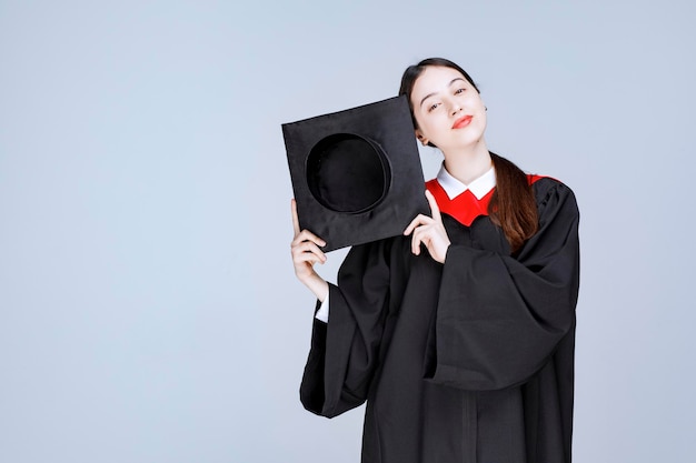 Joven estudiante en bata mostrando su gorro de graduación. Foto de alta calidad