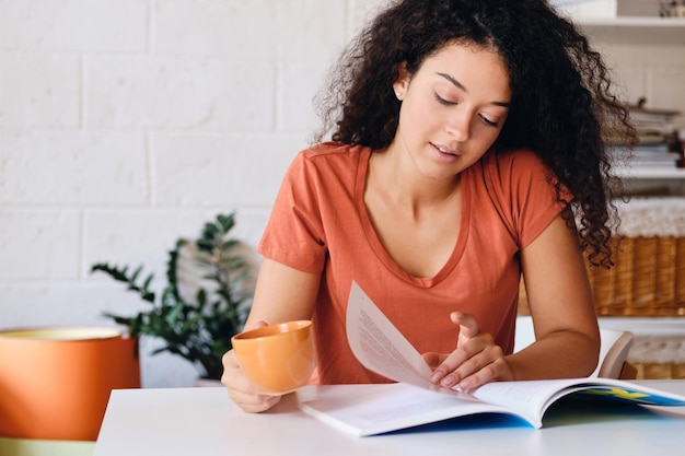 Joven estudiante bonita con cabello rizado oscuro sentada en la mesa con una taza de café naranja en la mano leyendo un libro soñadoramente en un hogar acogedor