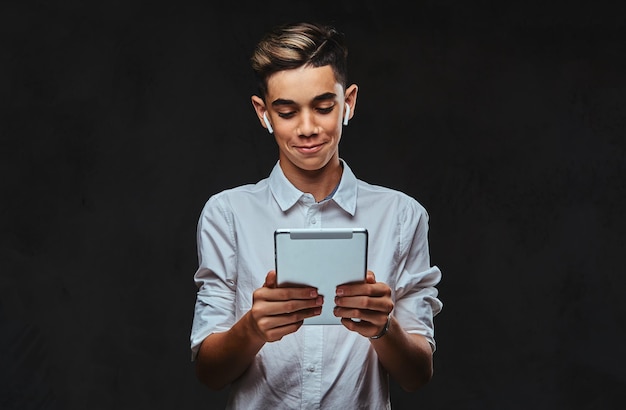 Joven estudiante con cabello elegante vestido con una camisa blanca usa auriculares inalámbricos y sostiene una tableta.