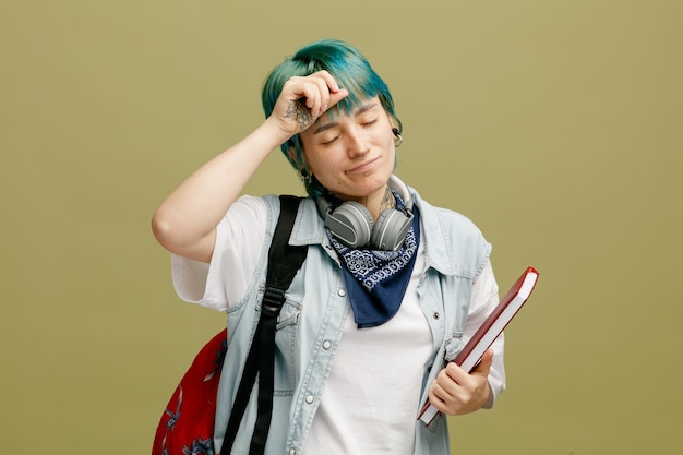 Joven estudiante cansada usando audífonos y pañuelo en el cuello y mochila sosteniendo un cuaderno tocando la cabeza con los ojos cerrados aislado en un fondo verde oliva