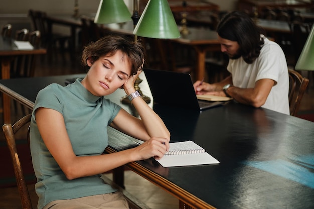 Joven estudiante casual cansada con bloc de notas durmiendo durante el estudio en la biblioteca de la universidad