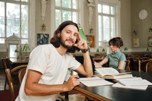 Joven estudiante casual que estudia con libros y conspectus mientras mira soñadoramente a la cámara en la biblioteca de la universidad