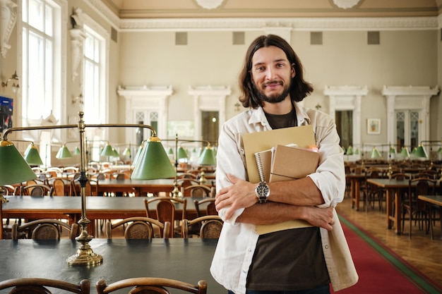 Foto gratuita joven estudiante casual sonriente con blocs de notas mirando felizmente a la cámara en la biblioteca de la universidad