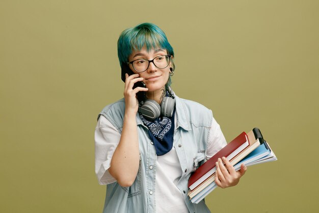 Joven estudiante complacida con anteojos, bandana y auriculares alrededor del cuello sosteniendo blocs de notas mirando a un lado mientras habla por teléfono aislado en un fondo verde oliva