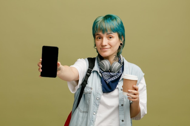 Joven estudiante complacida con auriculares y pañuelo en el cuello y mochila sosteniendo una taza de café de papel mirando a la cámara estirando el teléfono móvil hacia la cámara