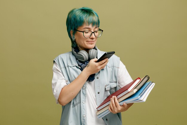 Una joven estudiante complacida con gafas, bandana y auriculares alrededor del cuello sosteniendo blocs de notas tomándoles una foto con un teléfono móvil aislado en un fondo verde oliva