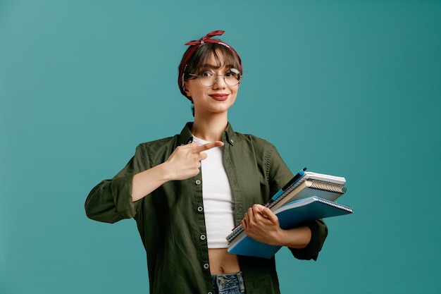 Una joven estudiante complacida con gafas de bandana sosteniendo grandes blocs de notas con un bolígrafo mirando a la cámara apuntando a los blocs de notas aislados en el fondo azul