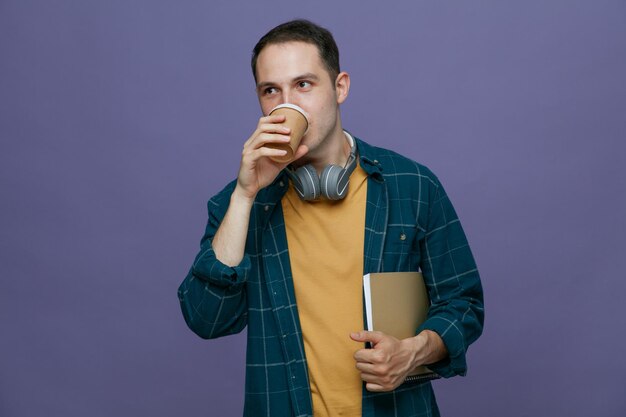 Joven estudiante concentrado usando audífonos alrededor del cuello sosteniendo un cuaderno bajo el brazo mirando al costado bebiendo café de una taza de café de papel aislada en un fondo morado