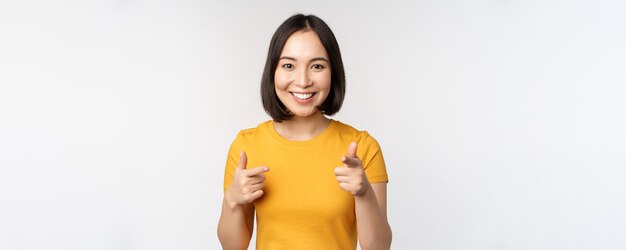 Joven estudiante coreana señalando con el dedo a la cámara y sonriendo felicitándote por elegir invitación