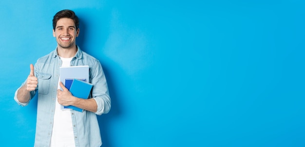 Foto gratuita joven estudiante con cuadernos que muestran el pulgar hacia arriba en señal de aprobación sonriendo satisfecho blue studio backgrou