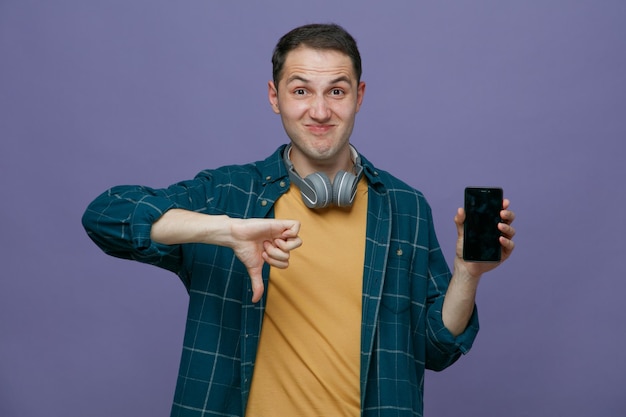Un joven estudiante descontento que usa audífonos alrededor del cuello mirando la cámara que muestra el teléfono móvil y el pulgar hacia abajo aislado en un fondo morado