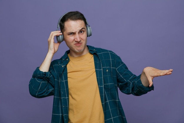 Foto gratuita un joven estudiante descontento usando audífonos agarrando audífonos mirando a la cámara haciendo un gesto de bajar la música aislado en un fondo morado