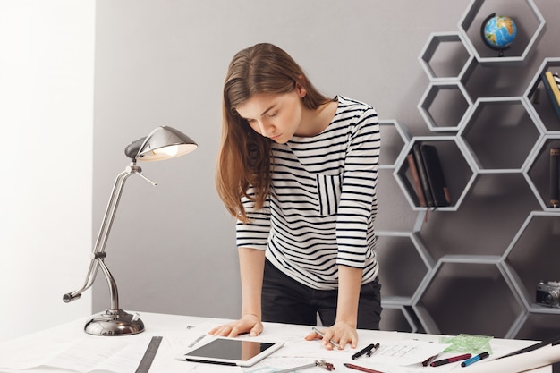 Foto gratuita joven estudiante de diseño serio serio con cabello oscuro en elegante traje casual de pie junto a la mesa, mirando en tableta digital, tratando de descubrir algunos detalles.