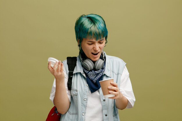 Joven estudiante enfadada con auriculares y pañuelo en el cuello y mochila sosteniendo una taza de café de papel y su gorra mirando hacia una taza aislada en un fondo verde oliva