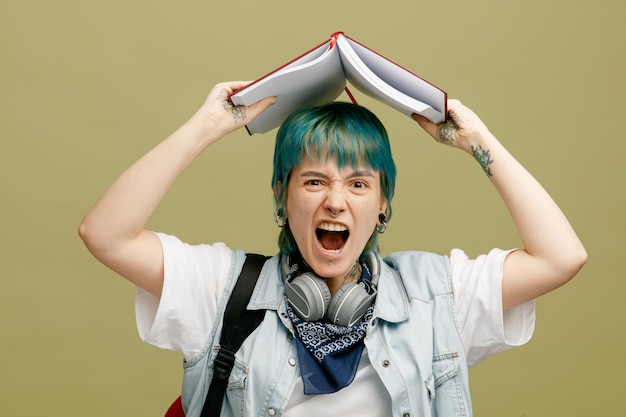 Joven estudiante estresada con auriculares y pañuelo en el cuello y mochila sosteniendo un cuaderno abierto sobre la cabeza mirando a la cámara gritando aislada en un fondo verde oliva
