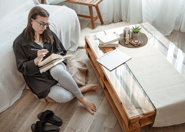 Una joven estudiante con gafas hace tareas en casa, sentada en un puf con un cuaderno en las manos.