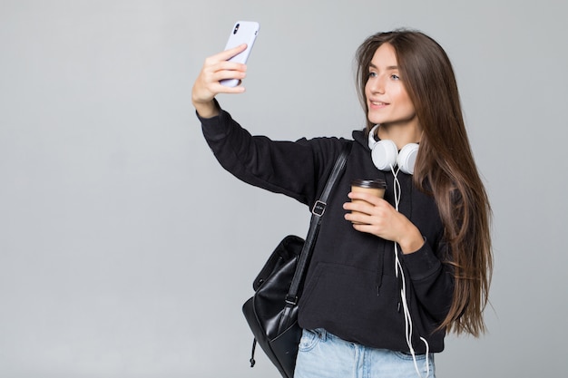 Joven estudiante hermosa chica con mochila hace selfie aislado en la pared blanca en estudio