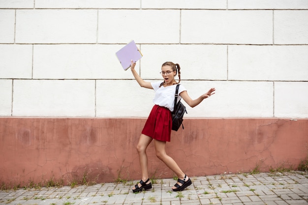 Foto gratuita joven estudiante hermosa en gafas caminando por la calle, sosteniendo carpetas.