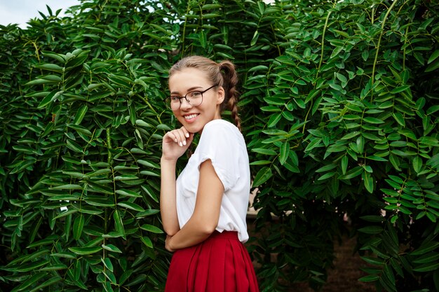 Joven estudiante hermosa en gafas sonriendo, posando sobre las hojas al aire libre.