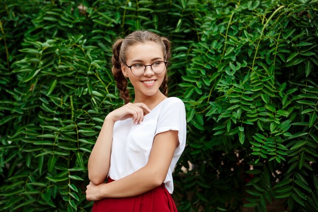 Joven estudiante hermosa en gafas sonriendo, posando sobre las hojas al aire libre.