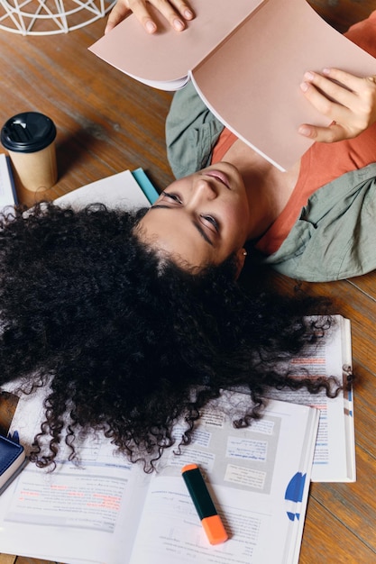 Joven estudiante hermosa con el pelo rizado oscuro tirado en el suelo con libros de texto alrededor estudiando soñadoramente en un hogar acogedor