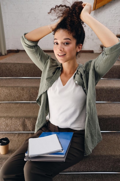 Joven estudiante hermosa sentada en las escaleras de la universidad con un cuaderno de rodillas y una taza de café para acercarse felizmente mirando a la cámara y atándose el pelo oscuro y rizado