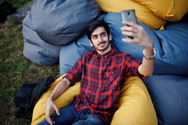 Joven estudiante indio con camisa a cuadros y jeans sentado y relajado en almohadas al aire libre y haciendo selfie