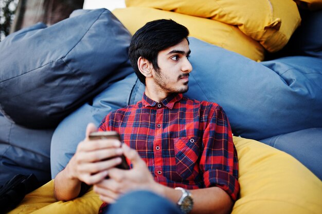 Joven estudiante indio con camisa a cuadros y jeans sentado y relajado en almohadas al aire libre Pasar tiempo con el teléfono móvil