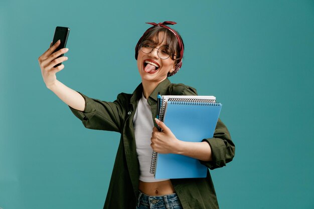 joven estudiante juguetona con gafas de bandana sosteniendo grandes blocs de notas con bolígrafo estirando el teléfono móvil mostrando la lengua tomando selfie aislado en fondo azul