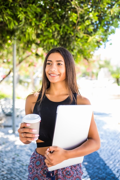 Joven estudiante latino hermosa con laptop y taza de café estudiando en el parque. La niña está caminando en el parque con una gran sonrisa y sostiene una computadora portátil.