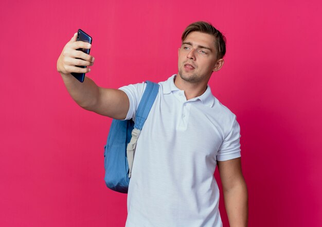 Joven estudiante masculino guapo con bolsa trasera tomar un selfie aislado en la pared rosa