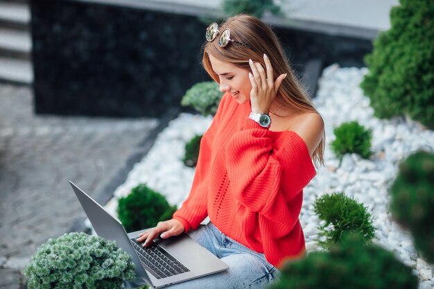 Joven estudiante, mujer rubia con camisa roja con laptop descansando en la calle urbana.