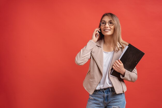 Joven estudiante o pasante sonriente en anteojos hablando por teléfono mientras está de pie con una carpeta